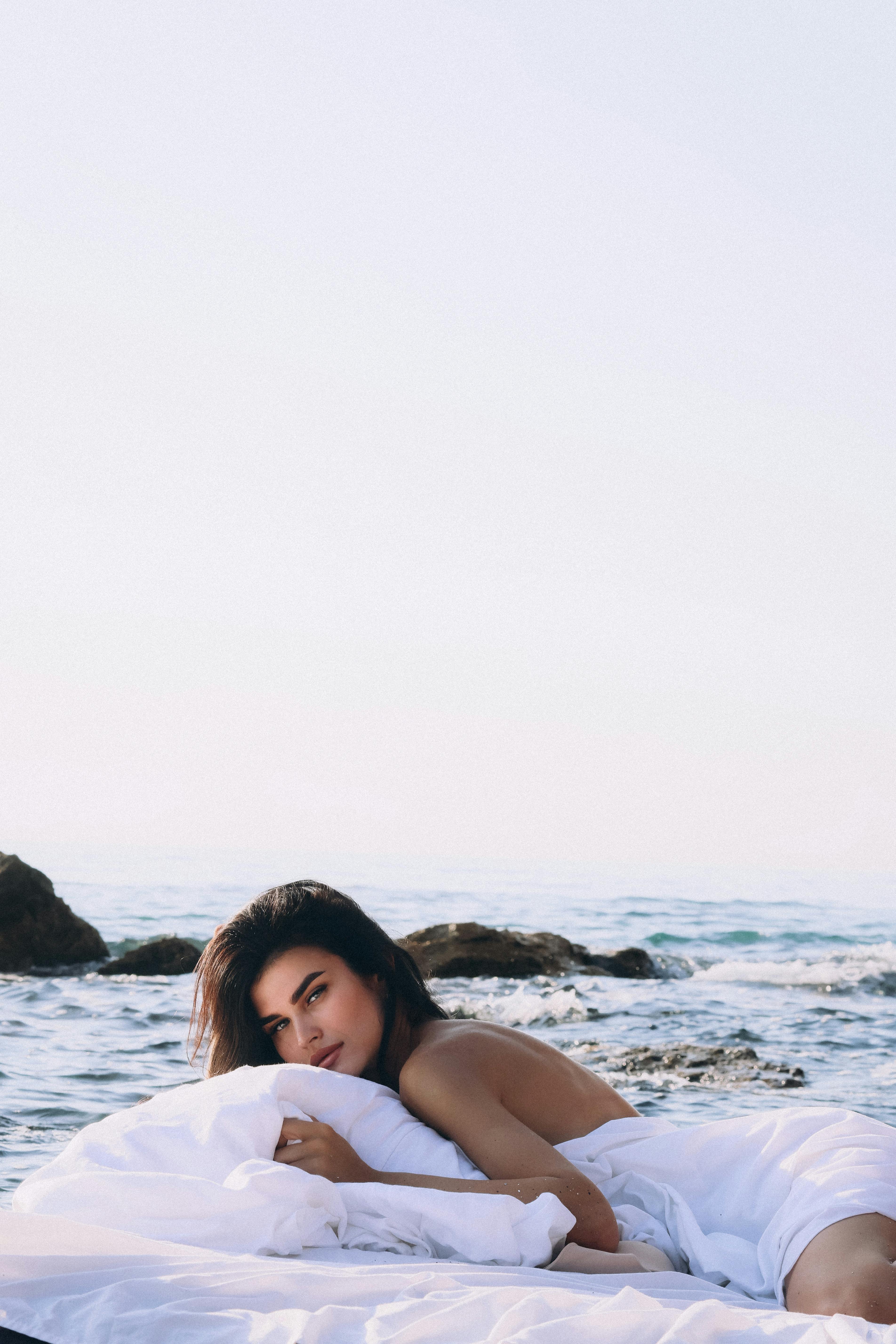 Person lying on a white blanket by the ocean with rocks in the background